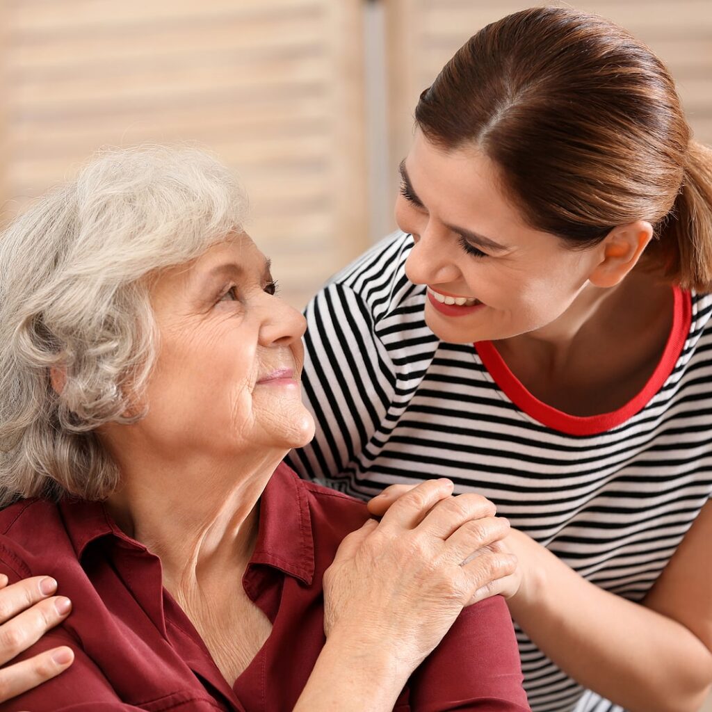 A younger woman in a striped shirt rests her hand on the shoulder of a smiling older woman