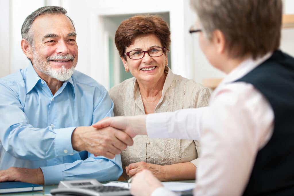 Elderly couple smiling while shaking hands with a professional across a desk in a bright office.